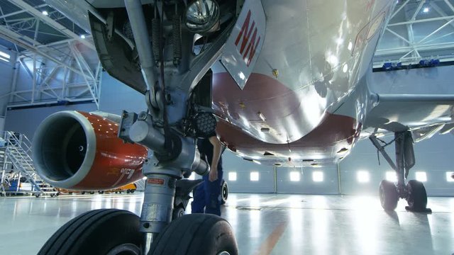 In a Hangar Aircraft Maintenance Engineer/ Technician/ Mechanic Visually Inspects Airplane's Chassis and Body/Fuselage walking Under It. Shot on RED EPIC-W 8K Helium Cinema Camera.