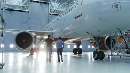 In a Hangar Aircraft Maintenance Engineer Shows  Technical Data on Tablet Computer to Airplane Technician. They Walk Alongside Clean Brand New Plane.Shot on RED EPIC-W 8K Helium Cinema Camera. - Powered by Adobe