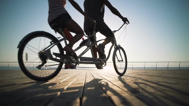 Portrait of a mixed race couple riding on tandem bicycle outdoors near the sea, throwing hat in the air slow motion
