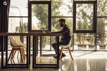 businessman using laptop while sitting at table in front of window in modern office