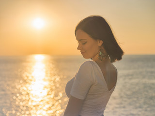 Portrait of young attractive woman standing in morning sunlight close to sea or ocean. Beautiful lady with short brunette hair and long earrings.