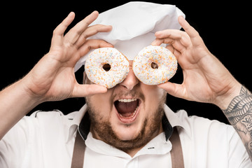 Excited chef in hat and apron looking through doughnuts isolated on black