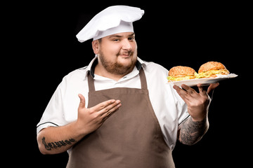 portrait of smiling cooker pointing at plate with burgers isolated on black