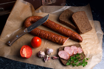 Slices of smoked sausage with spice, herbs and vegetables on the packaging paper. Vintage knife.