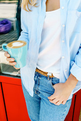 Hipster girl in jeans holding a cup of cappuccino near cafe. Beautiful lifestyle portrait