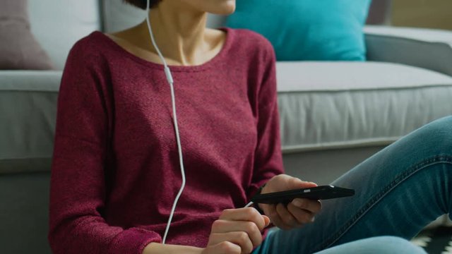 Young caucasian beautiful woman with short dark hair sitting on the floor relaxing listening to music using black cell phone and white earphone. Indoor.