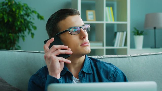 Handsome caucasian man in glasses with dark hair at home, sitting on couch talking on the smart phone and using his laptop.