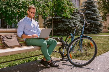 Fototapeta premium Closeup of businessman hands typing on laptop while coffee breaks in park. Casual businessman sitting on a bench and working, next the bench rests a bike.