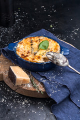 Lasagna in baking dish on an old chopping board, basil, old spoon, blue napkin, parmesan, rosemary on a dark metal table. Close-up. Italian food.