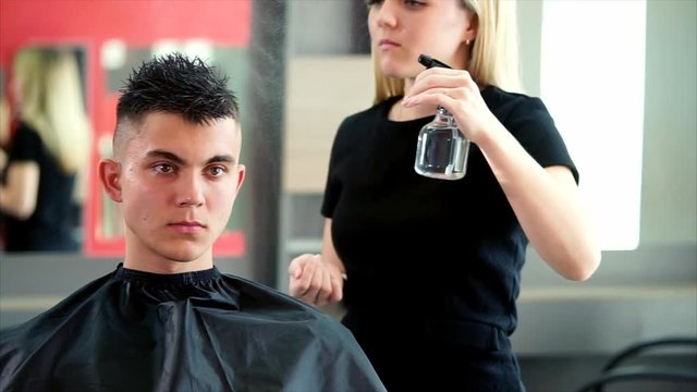 A young and professional hair stylist who works in a hair salon sprinkles water from an atomizer on the visitor's hair in order to make a fashionable hairstyle.