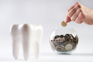 Hand put money in piggy bank filled with coins and white healthy tooth isolated on white background...