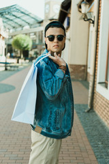 Fototapeta premium young stylish man in sunglasses holding shopping bag and standing on the street