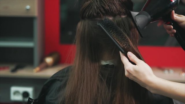 close up shot of the hairdresser's hands, who dries dark and long hair with a hair dryer and comb, the beauty salon client is waiting for the completion of the image change