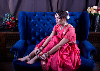 The girl in a scarlet sari and Indian jewelery corrects anklet sitting on a soft blue sofa surrounded by vases with flowers
