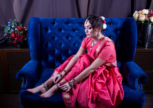 The girl in a scarlet sari and Indian jewelery corrects anklet sitting on a soft blue sofa surrounded by vases with flowers
