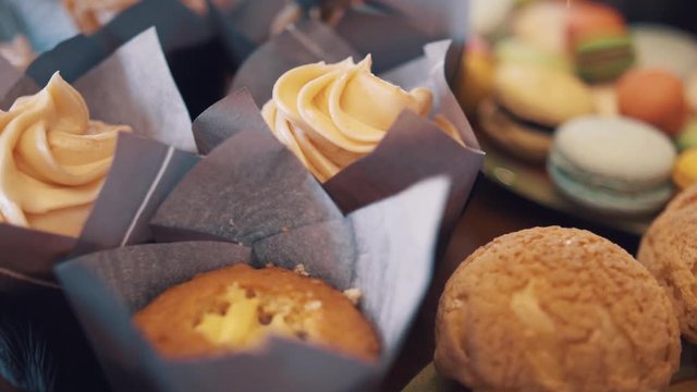 Pastry cook makes creme swirl on stuffed muffins wraped in purple paper on table with sweet foods, close up
