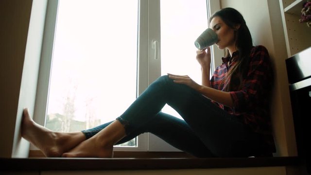 Beautiful young woman drinking coffe, tea while sitting on windowsill at home. Woman dreaming and smiling.