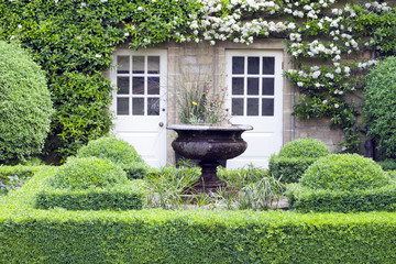 Flower vase in green topiary garden in front of a stone English country house, with two patio doors...
