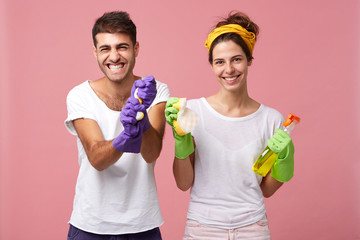 Positive couple doing home chores: stylish man in protective gloves holding sponge and smiling housewife holding rag with spray standing together isolated over pink wall. Housework, cleaning concept