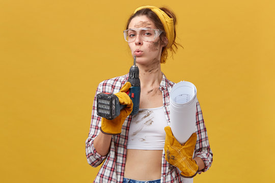 Self-confident female builder wearing goggles, white top and checkered shirt, protective gloves holding drill and papers being dirty after hard work isolated over yellow background. Maintenance