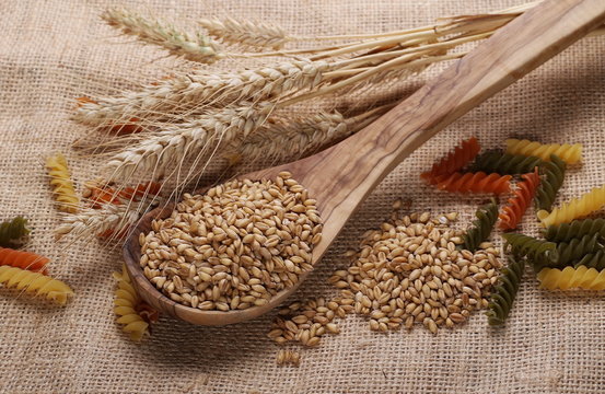 Ears of wheat, seeds, spiral macaroni and wooden spoon on linen, jute background, texture