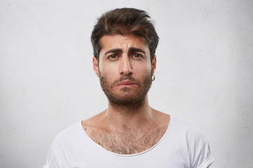Fototapeta premium Stressed bearded male having sad expression wearing earring and white T-shirt curving his lip knowing sad news. Bemused male isolated over white background. People and negative emotions concept