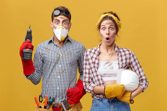 Young service workers doing maintenance: male with protective mask, eyewear, gloves and belt with tools holding drill and woman in checkered casual shirt holding hardhat having astonished looks