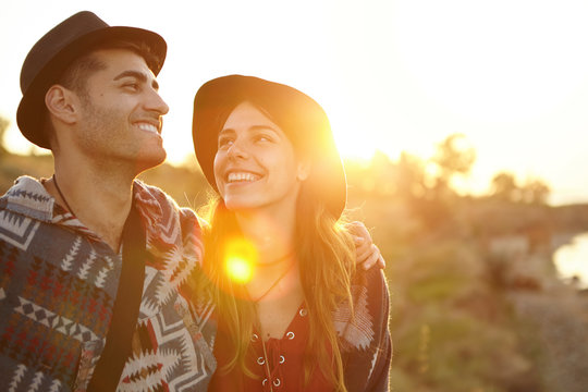 Two People In Love Enjoying Sunshine Embracing Each Other Looking With Love Having Eyes Full Of Happiness Standing Against Green Nature Background. Couple Having Walk Through Meadow Admiring Nature