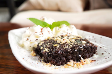 Sliced homemade brownie on white plate. Served with whipping cream and mint leaf decorate. Over wooden table.