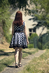 Young woman walking on village way. Woman in spotted dress and red hair. Sunny and positive rural scene with woman walking away from camera.