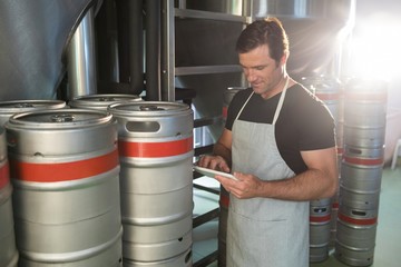 Worker using digital tablet on notepad while standing by kegs