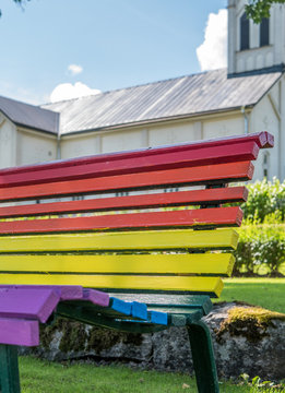 Rainbow colors on bench, church in background.