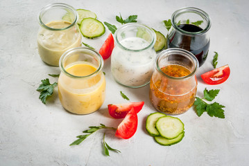 Set of dressings for salad: sauce vinaigrette, mustard, mayonnaise or ranch, balsamic or soy, basil with yogurt. Dark white concrete table, with greenery, vegetables for salad. Copy space