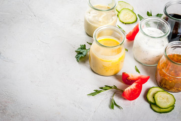 Set of dressings for salad: sauce vinaigrette, mustard, mayonnaise or ranch, balsamic or soy, basil with yogurt. Dark white concrete table, with greenery, vegetables for salad. Copy space