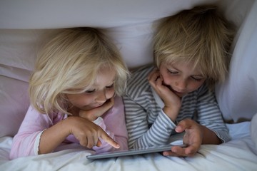 Siblings using digital tablet under blanket on bed in bedroom