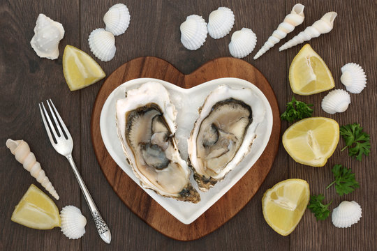 Oysters on crushed ice on a heart shaped plate on a maple wood board over old oak background with lemon fruit, parsley, sea shells and an antique fork.