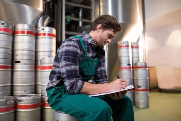 Worker writing on paper while sitting on keg at warehouse - Powered by Adobe