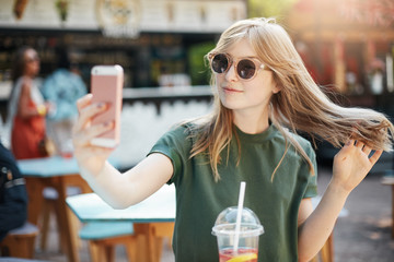 Blonde freckled girl making a selfie or chatting via video call with her friends sitting in park on a summer day.