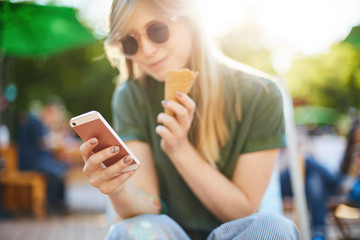 Woman eating icecream using smartphone. Portrait of happy girl with ice cream browsing through social media or messaging her friends enjoying summer in the city park wearing shades. Focus on phone.