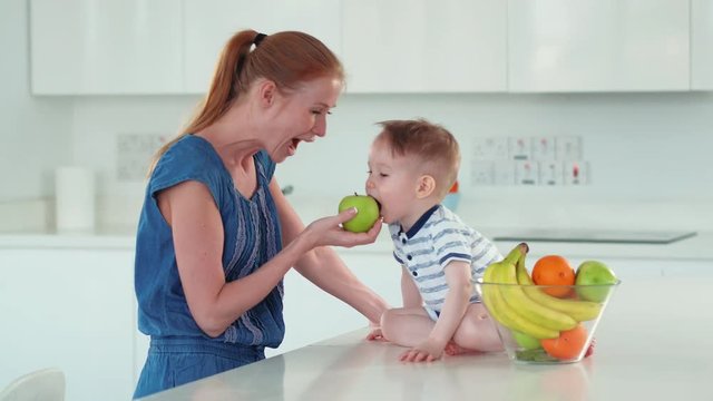 Baby boy with his mother on the white kitchen playing with colorful fruits