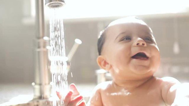 Baby in Sink Bathing and Smiling Looking Away. a stream of water pouring out of the faucet tap and baby looks off camera smiling and laughing then turns interest towards water in slow motion

