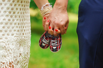 Future parents holding hands and a pair of little shoes overnature green background. Concept of...