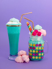 Delicious homemade extreme milkshake in a jar and a cup of glass, with a blackberry candy over a milk foam and a rainbow candy on top with a plastic straw in a purple background