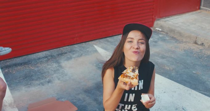 Two stylish teen girls with a longboard eating pizza outside, red metal wall urban background. 4K UHD