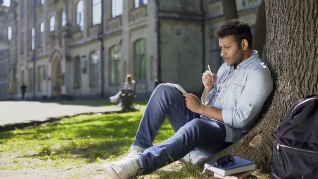 Multiracial male sitting on grass under tree, writing in notebook, creative idea