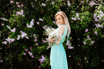 Obraz na płótnie Canvas Blonde girl with bouquet of wild flowers outdoors A model in a turquoise dress against a background of a lilac bush.