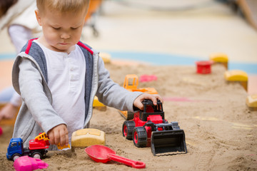 Portrait of cute toddler boy sitting on the ground and playing with toy tractor and sand in the park. Child walking outdoors. Lifestyle.