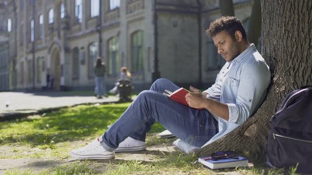 Male university student sitting under tree, reading book, memorizing information