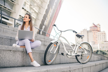 Beautiful woman office worker at lunch break working on laptop. A young woman is sitting on the steps, next to the bike.
