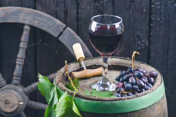 Glass and bottle of wine next to wooden barrel. Burnt, black wooden background. Vintage. Copyspace for a text. Grapes and green vine. 